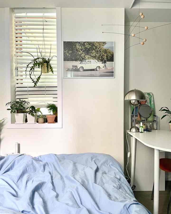Light neutral white bedroom featuring white walls, blinds, abundant plants, and a light blue bed with tidy desk creating cozy atmosphere
