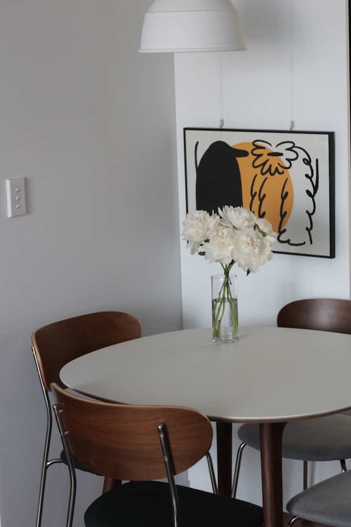 A white-toned dining room with a round table featuring a glass vase with white flowers, surrounded by wooden dining chairs creating a fresh atmosphere