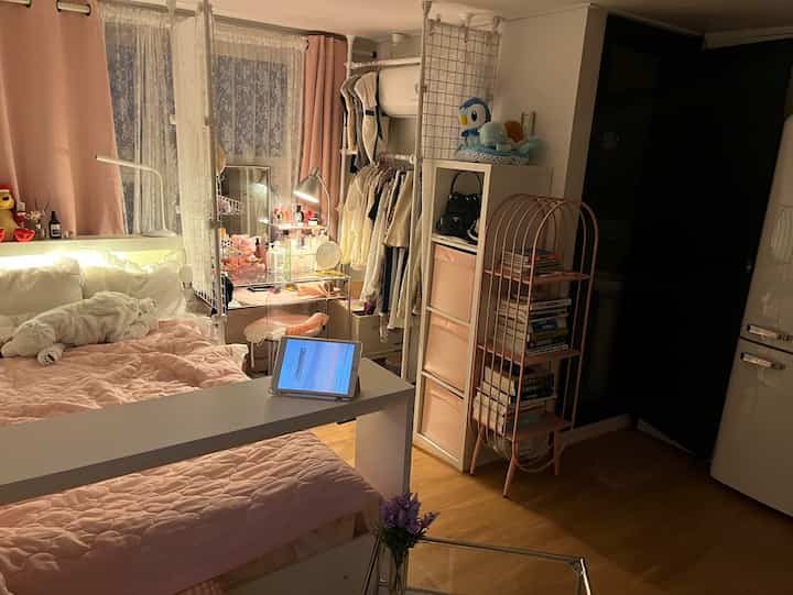 ピンクとホワイトの色調の寝室、ベッド、デスク、ハンガーラックが配置された印象的なコンパクトな空間