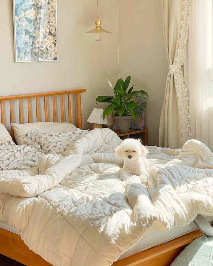 暖かいベージュトーンの寝室、木製ベッドと白い寝具セット、窓際のカーテンと緑の植物が調和した雰囲気のあるインテリア