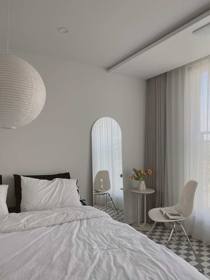 White and beige toned bedroom featuring a large bed, pillows, arched full-length mirror, side table, and chairs with a clean, simple atmosphere