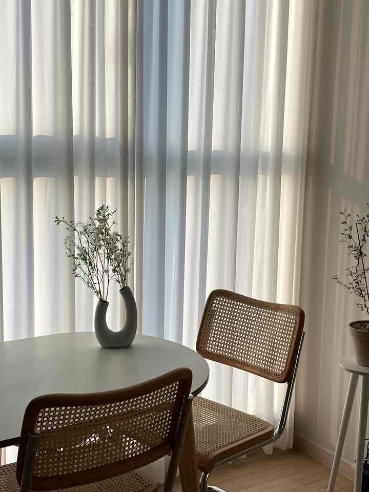 明るいホワイトカーテンの前にベージュの木目調ダイニングテーブルとラタンチェアが配置されたナチュラルなダイニングルーム