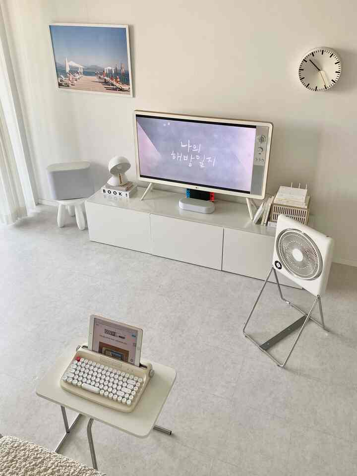 White-toned living room featuring a clean TV stand, wall clock, and minimalist table lamp with a simple modern interior
