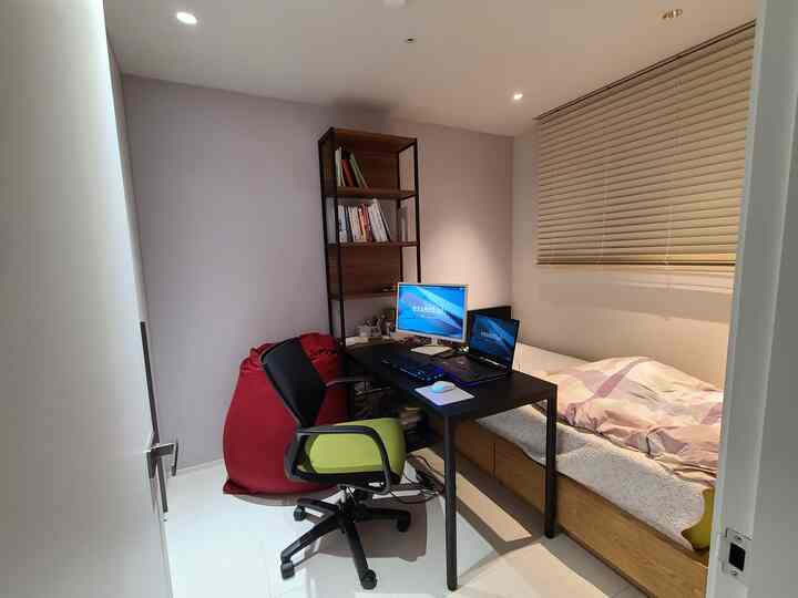 A compact bedroom with white walls and floor, featuring a black desk, green office chair, red bean bag, and wooden bookshelf, creating a cozy gaming and study space