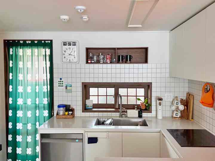 White and green toned kitchen featuring natural wood window frame and shelves, Twemco flip clock, and patterned curtain for a cozy kitchen interior