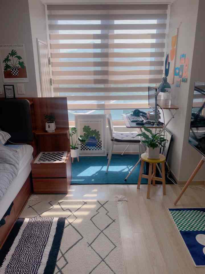 白と木目調が調和したワンルーム、ベッドとデスク、ブラインドが配置されたシンプルでモダンな在宅勤務スペース