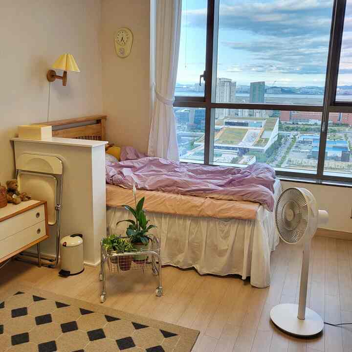 Bright natural-tone studio bedroom featuring white curtains, a bed, fan, and rug in a cozy space