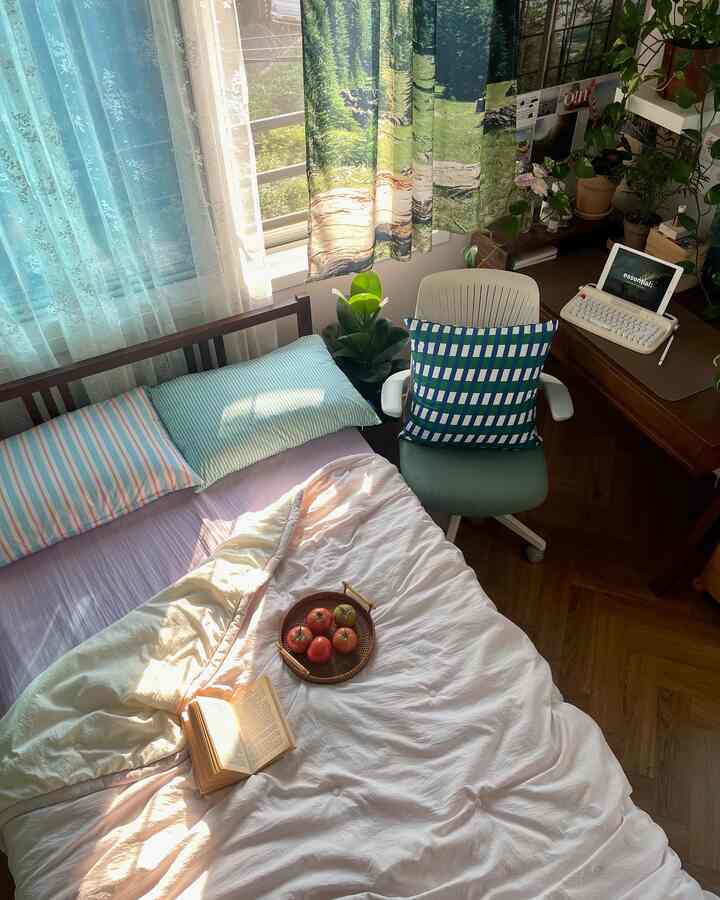 Cozy bedroom with green plants and curtains featuring forest pattern, desk and chair with blue-green cushion, white bedding