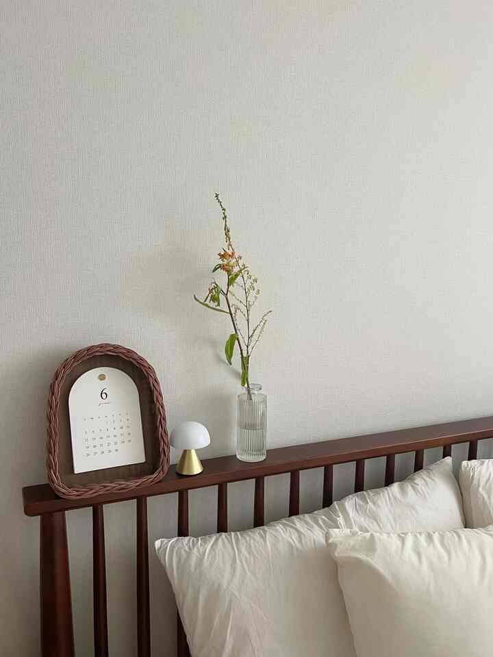 White and brown toned bedroom featuring a wooden bed frame with a small table lamp, a vase with a single flower, and a framed calendar, creating a minimalist peaceful space