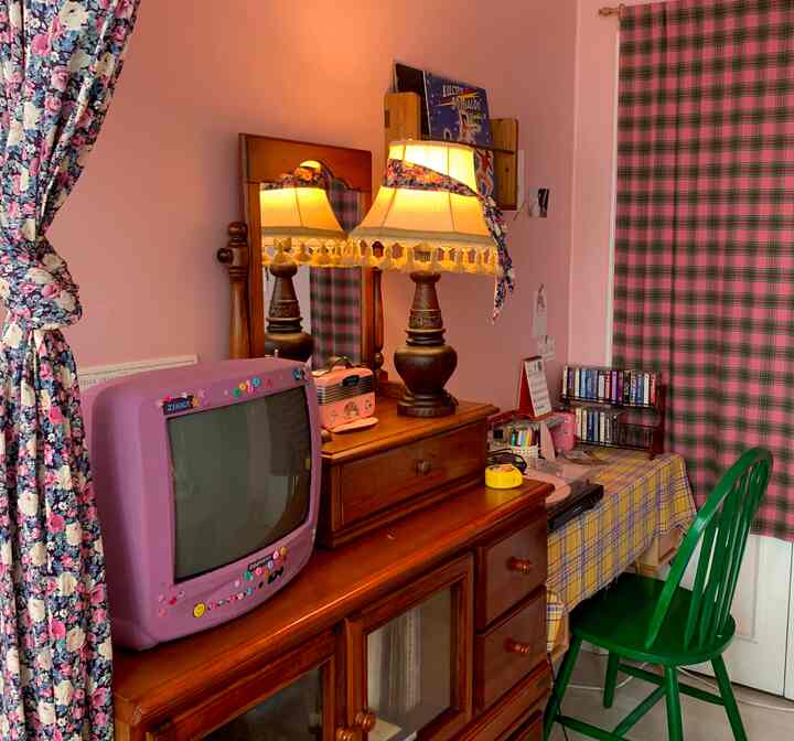 A retro-styled living room with pink-toned walls and brown vintage wooden furniture, adorned with vintage accessories and floral curtains, creating a cozy atmosphere