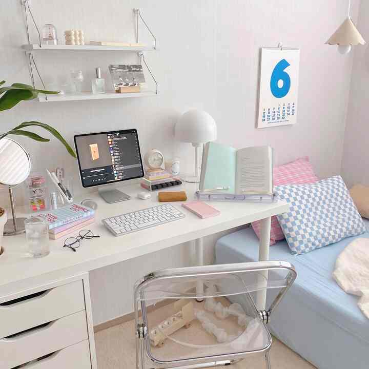 Compact home office and bedroom with white and pastel blue tones, featuring a desk, transparent chair, wall shelves, and pillows in a tidy setting