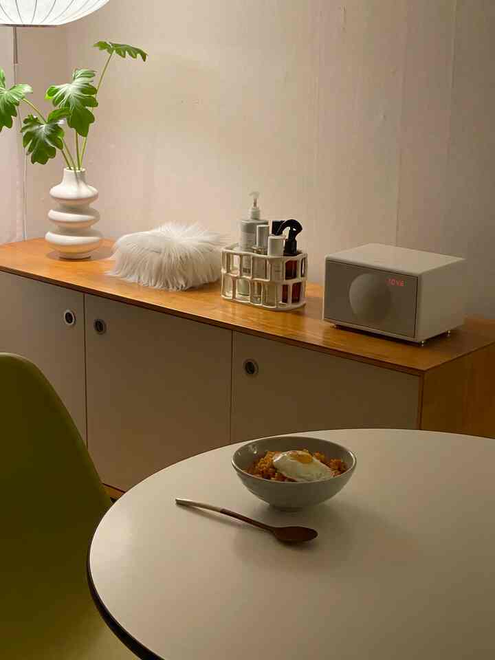 白と茶色のトーンのダイニングルーム、丸いダイニングテーブルと青い椅子、木製キャビネットの上に花瓶とオーディオが配置されたミニマルな空間