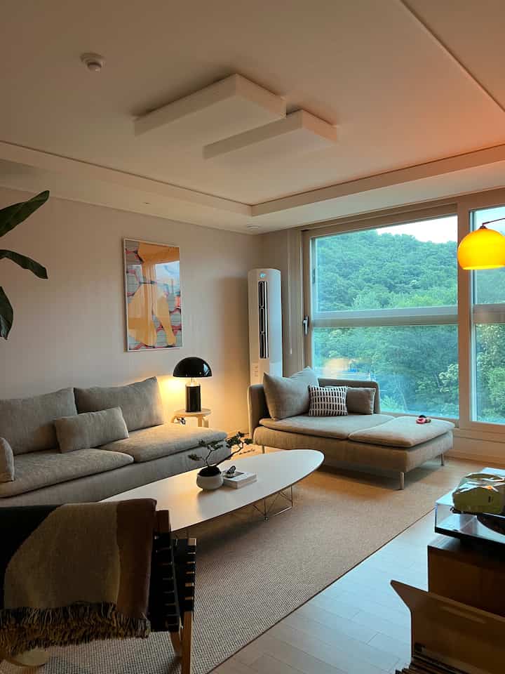 Beige and white toned living room featuring natural modern sofa, rug, and large window showcasing lush greenery outside, creating a cozy atmosphere