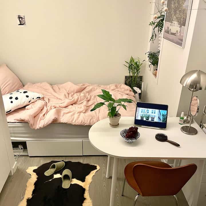 Cozy studio bedroom with soft pink bedding, white furniture, and green plants, presenting a compact and inviting atmosphere