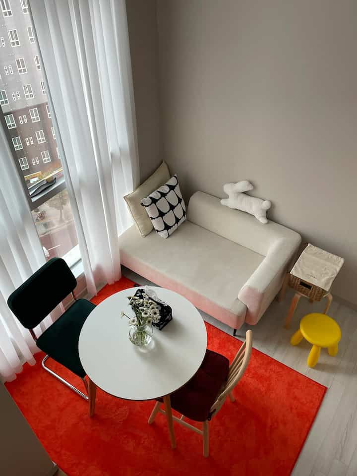 Bright white and orange-toned living room featuring a beige sofa, round coffee table, and yellow stool in a simple, cozy interior