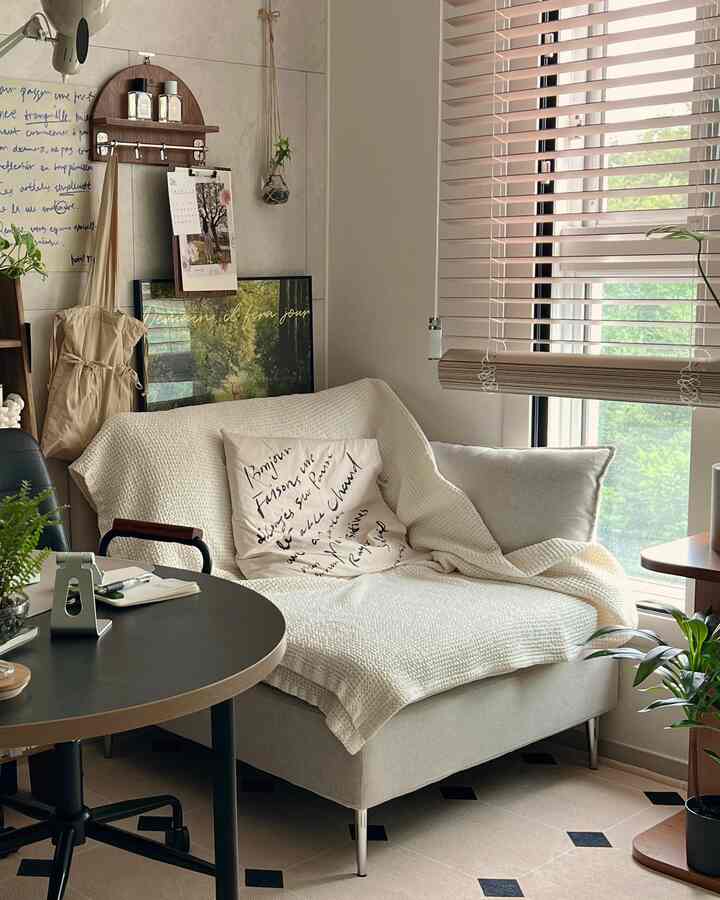 A natural-toned living room in white and beige, featuring a sofa, office desk, and blinds by a window