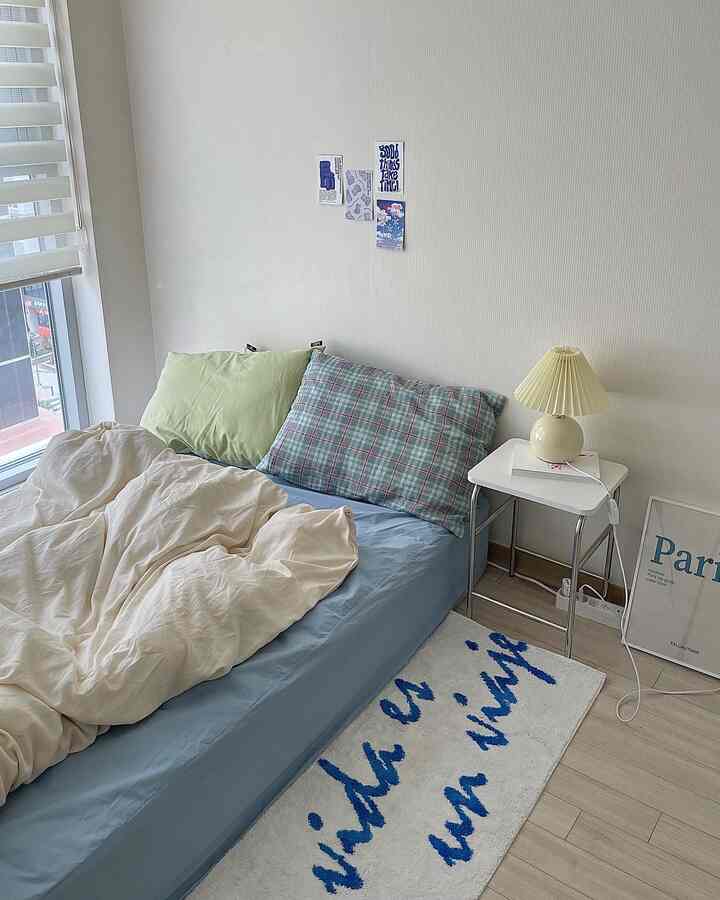 A white and blue toned bedroom featuring a blue mattress and pillows, white nightstand with a table lamp in a cozy space.