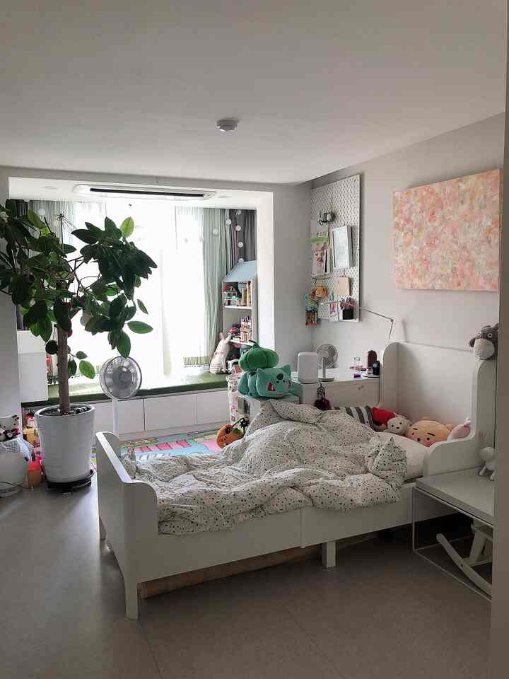Modern kids' room blending white and natural tones featuring a white bed, bookshelf, fan, and large indoor plant