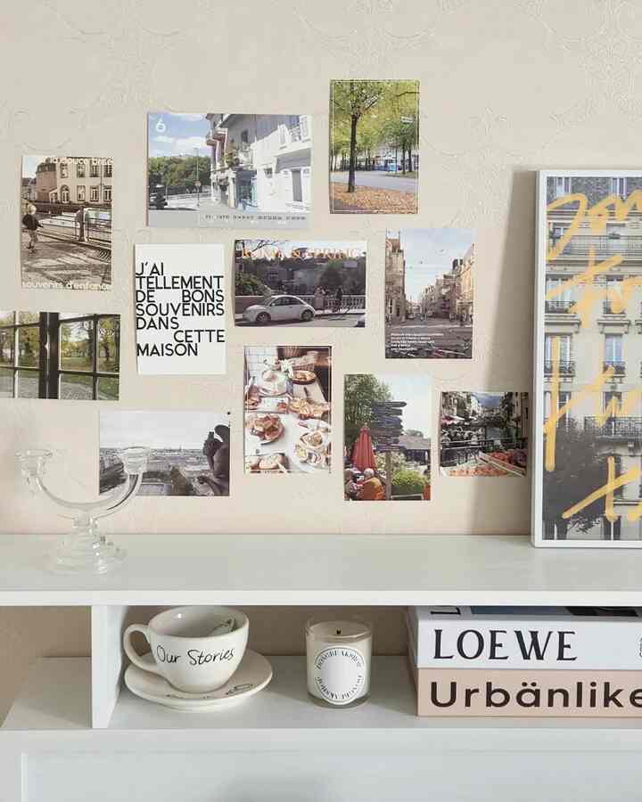 A cozy bedroom with soft beige and white tones featuring European-style postcards and posters on the wall, white shelf below with cup and candle accents