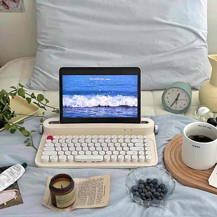 淡いブルーの寝室、中央にレトロなキーボードとタブレットが置かれ、横に置時計とマグカップのある落ち着いたおうちカフェ風インテリア