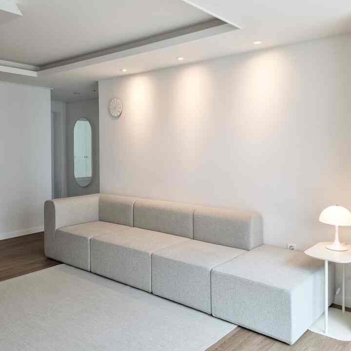 White and gray toned living room featuring a modern sofa, side table with lamp, and a wall clock in a simple layout