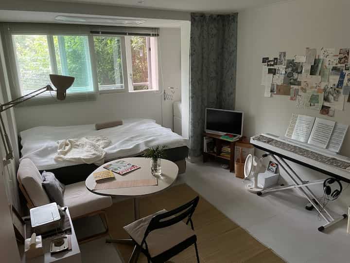 ウッドトーンとナチュラルカラーのワンルーム寝室、丸テーブルとデジタルピアノが配置された落ち着いた空間