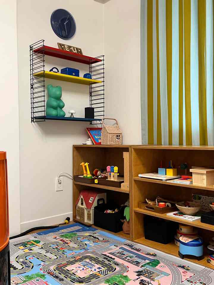 白い壁と木目調の収納棚が配置された子供部屋、多彩なおもちゃとベージュのストライプカーテンが調和する印象的な空間