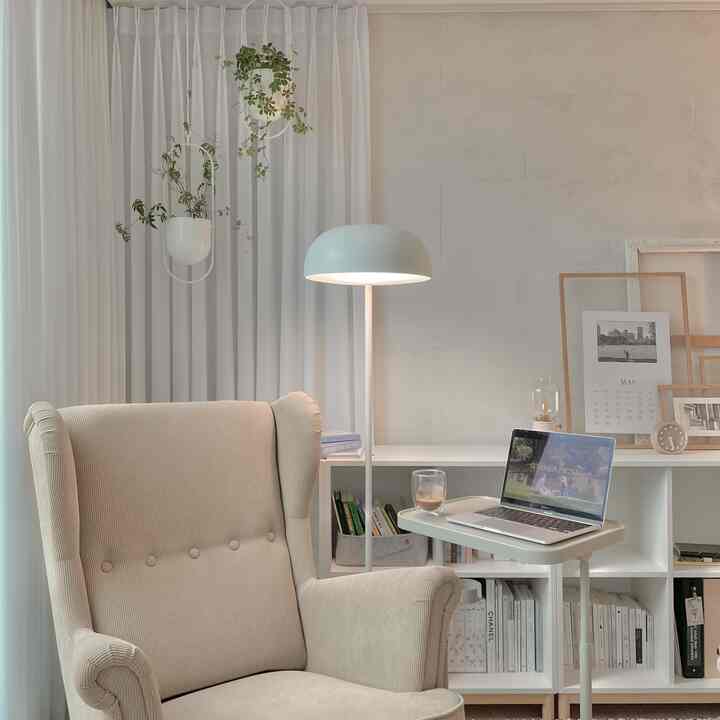 White and beige toned study-style living room featuring an armchair, floor lamp, bookshelf, and laptop, creating a cozy ambiance
