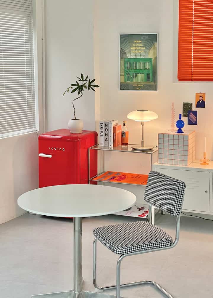 A red and white toned studio living room featuring a red mini refrigerator, round table, and checkered chair in a mid-century modern style