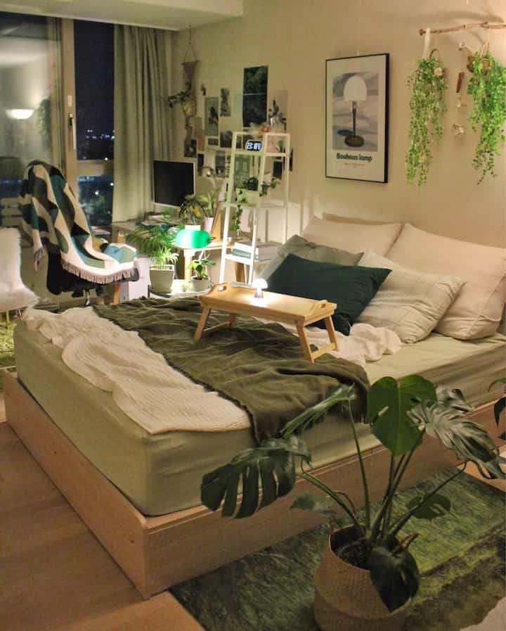 Green and natural toned studio bedroom featuring a bed and various plants with cozy ambiance