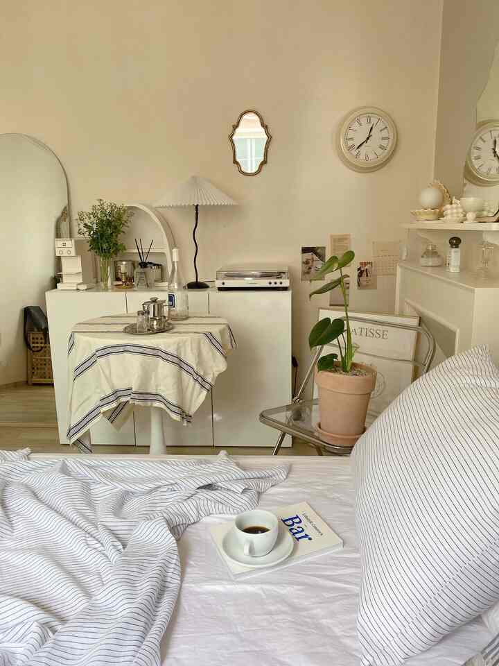 淡いベージュとホワイトの色調の寝室、整えられたベッドと小さなテーブル、植物と時計が配置された雰囲気のあるインテリア