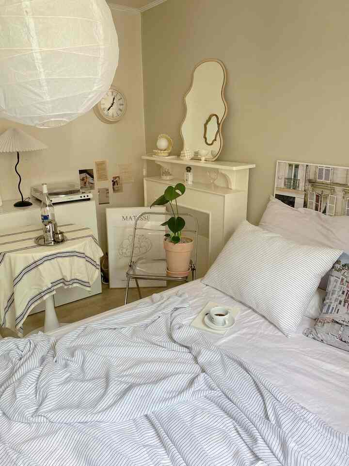 A minimal bedroom with soft beige walls and white bedding, featuring a transparent chair with plant and a unique wall mirror