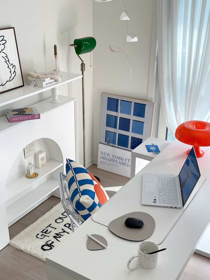 A modern home office with white backgrounds and blue tones, featuring a clean desk, floor lamp, colorful cushion, and a striking poster