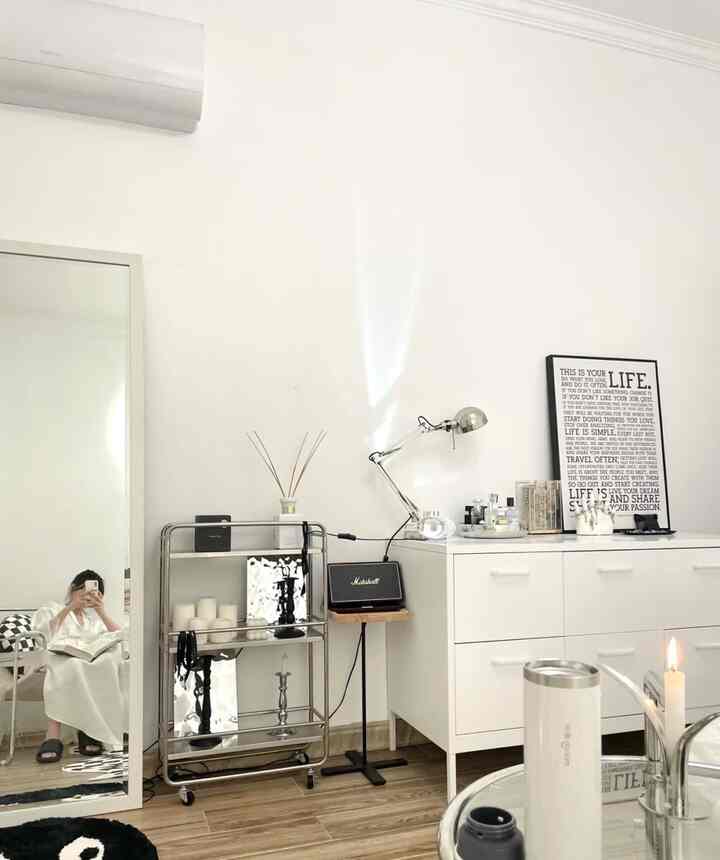 Bright white-toned studio bedroom featuring minimal furniture, black and white wall art, and a multi-tier storage cart in a neat arrangement