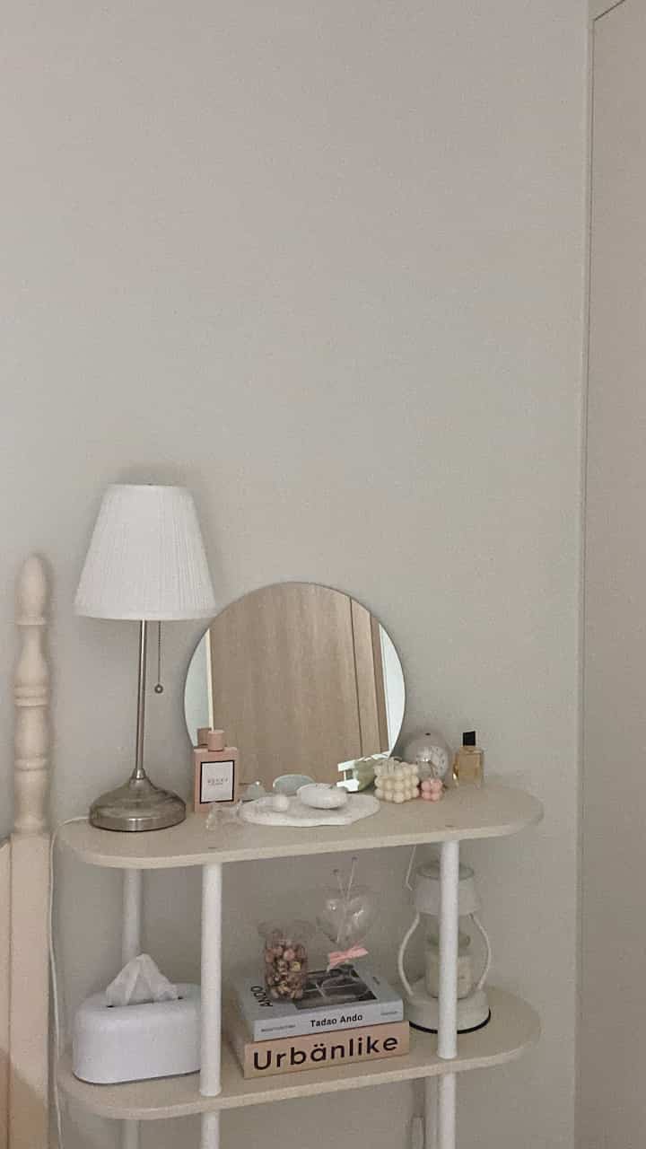 Ivory and beige toned bedroom corner with a round mirror and table lamp on a simple nightstand space