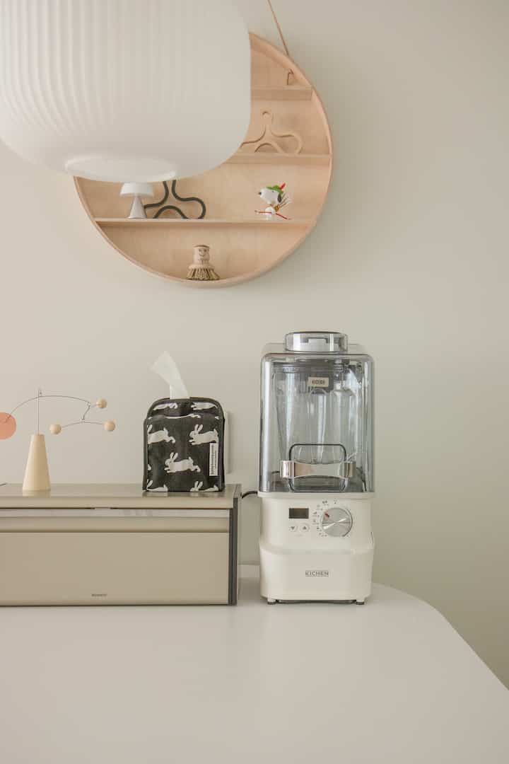 ベージュの収納と丸い木製の棚がある白いトーンのキッチン、中央にキチェンの真空ブレンダーが置かれたミニマルな空間