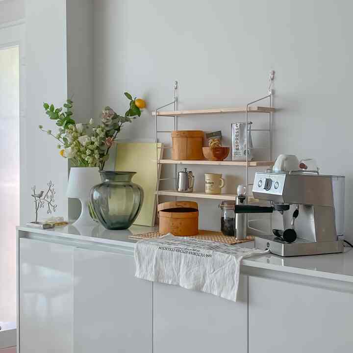 白と木目調のリビングのおうちカフェ、コーヒーマシンや収納棚、花瓶が配置された雰囲気のあるインテリア