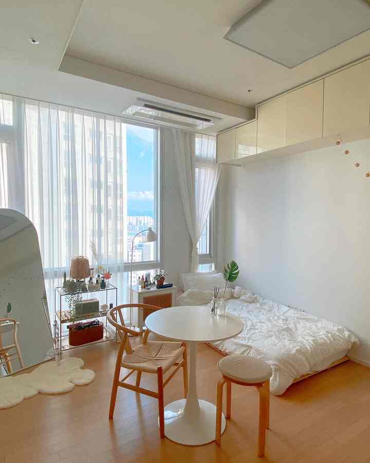 Bright white and wood-tone high-rise studio apartment featuring a round table and a cozy bed in a natural setting