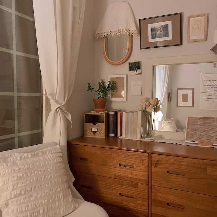 Cozy small bedroom featuring brown wooden dresser and ivory blackout curtain, vintage wall decor, and vanity mirror in a nature-inspired setup