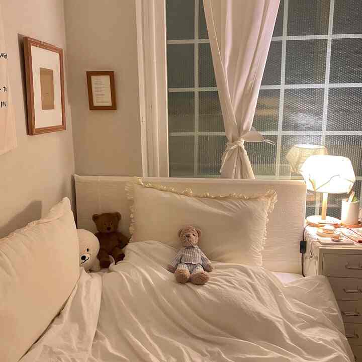 Cozy bedroom in warm white tones featuring white blackout curtains, brown picture frames, and teddy bears on the bed