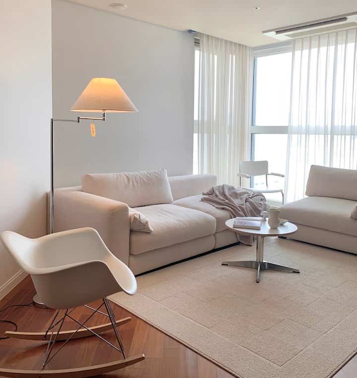 Soft white-toned living room featuring two large sofas, a unique rocking armchair, floor lamp, and curtains creating a modern atmosphere