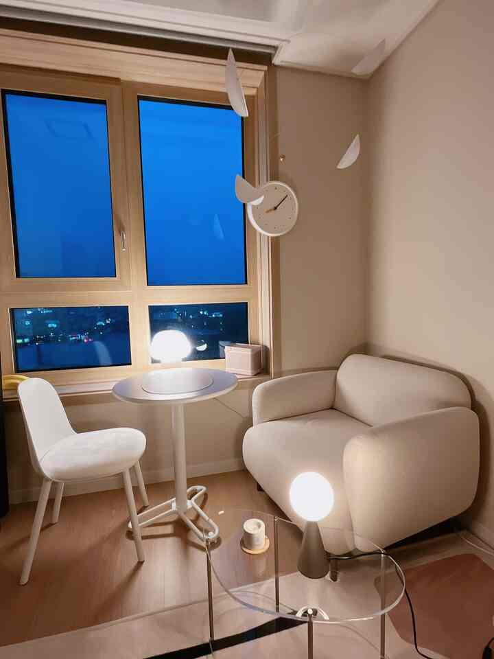 アイボリー調のワンルームのリビング、窓際に丸テーブルとソファ、チェア、照明が配置された温かみのある空間