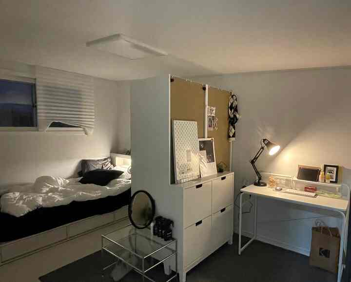 Compact single bedroom in white and black tones, featuring a bed and storage cabinet dividing the space, with a home office desk arranged on the right, reflecting simple modern design.
