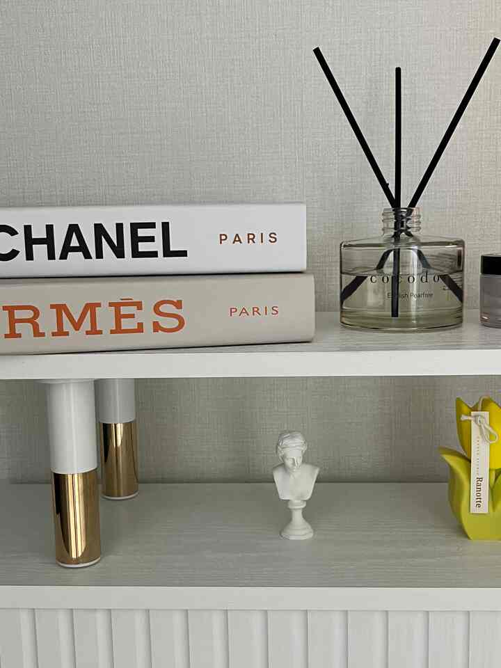 A white bookshelf with gold accents displaying a diffuser and decorative objets in a clean, minimalist interior setup