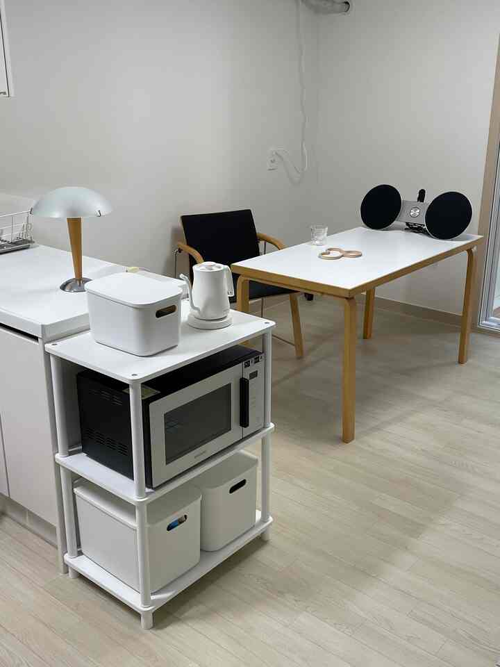 ホワイトとウッドトーンが調和したシンプルなリビングとキッチンスペース、ヴィンテージ風のアームチェアとシンプルなテーブルランプが配された若者向けの住宅。