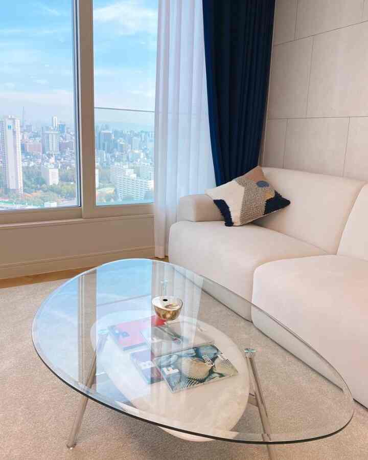 Bright and spacious living room featuring a transparent glass coffee table and ivory sofa, with a large window showcasing a sky view