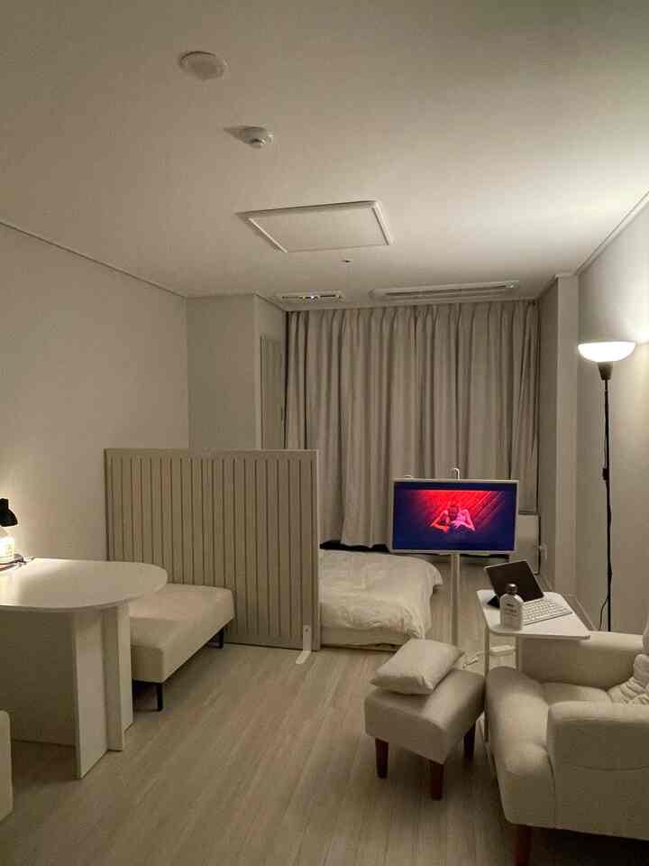 白を基調としたワンルーム寝室、リクライナーソファとスツール、テレビスタンドが配置されたホテルライクな落ち着きのあるインテリア