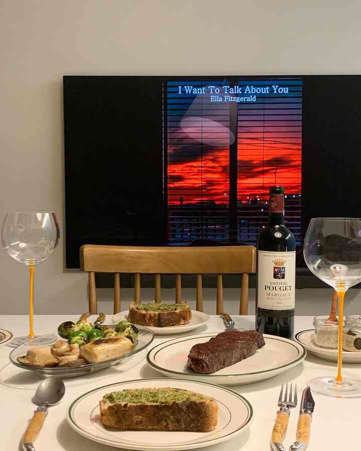 A simple home cafe space in a living room with wood-tone chair, wine glasses, steak on a dining table, and a vivid red sunset on TV