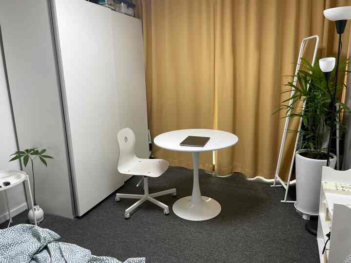 Compact bedroom in white and yellow tones featuring a round table with chair, curtains, floor lamp, and plants arranged cozily