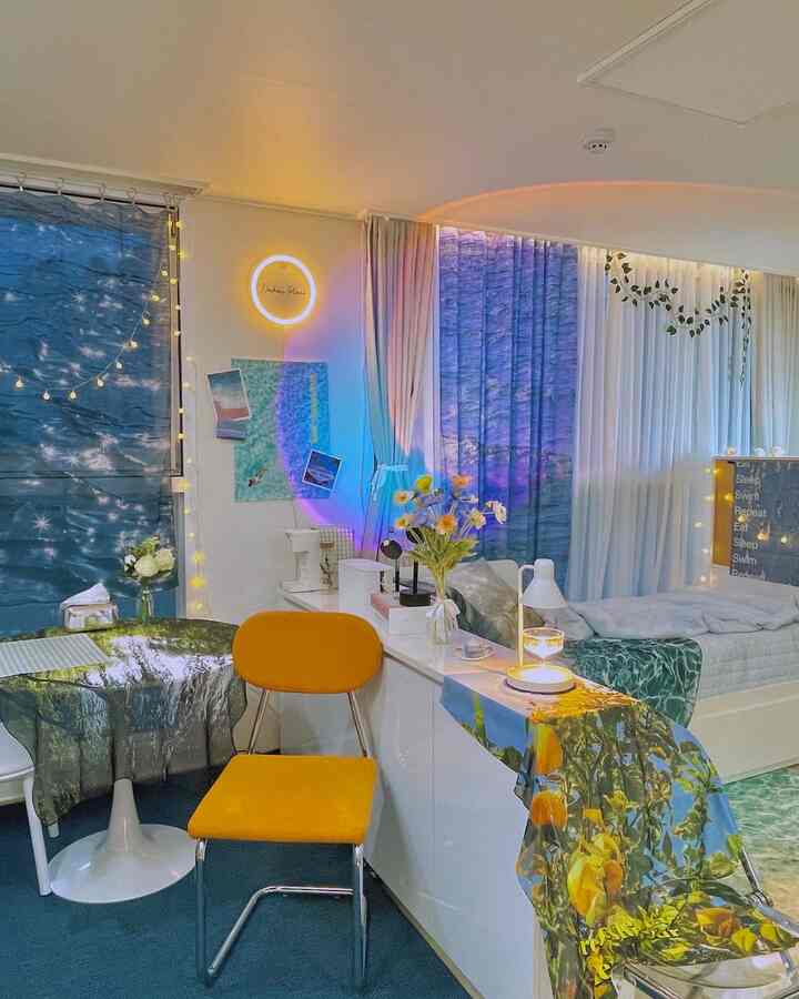 Studio bedroom with blue curtains and yellow dining chair, featuring lighting and floral decor for a lively ambiance